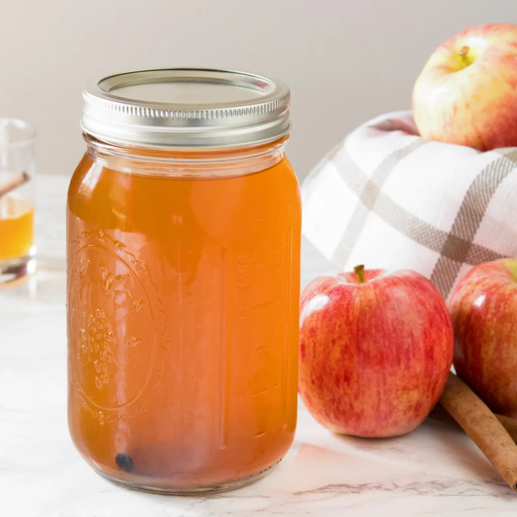 Homemade apple pie moonshine in a mason jar with apples, cinnamon sticks, and spices on a rustic wooden table.